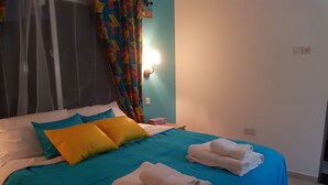 A neatly made bed with blue and white bedding, a colorful curtain, and a wall-mounted light. - Camara Gardens Apartments with Pool (Brufut)