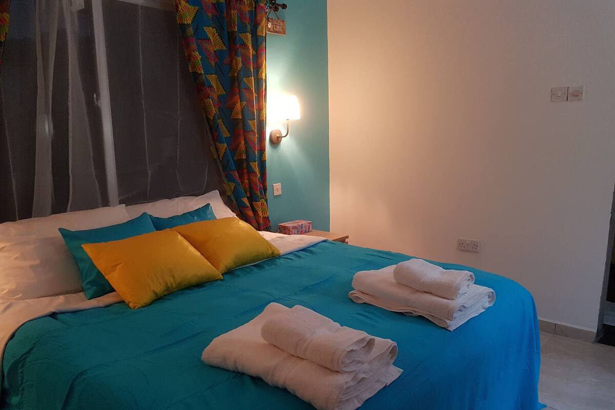 A neatly made bed with blue and white bedding, a colorful curtain, and a wall-mounted light.