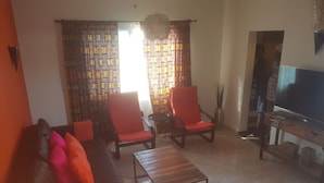 A living room with a sofa, two red armchairs, a coffee table, and a TV on a wooden stand. - Camara Gardens Apartments with Pool (Brufut)