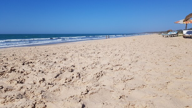 A beach with footprints leading to the water, a few people in the distance, and beach umbrellas on the right. - Camara Gardens Apartments with Pool (Brufut)