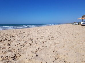 A beach with footprints leading to the water, a few people in the distance, and beach umbrellas on the right. - Camara Gardens Apartments with Pool (Brufut)
