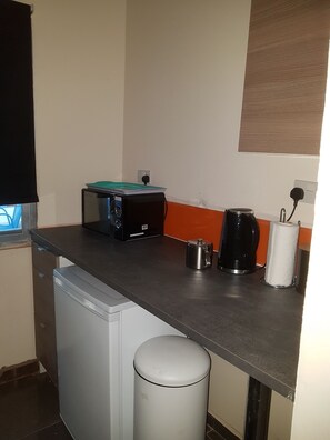 A compact kitchenette with a small refrigerator, a coffee maker, a kettle, and a trash bin.