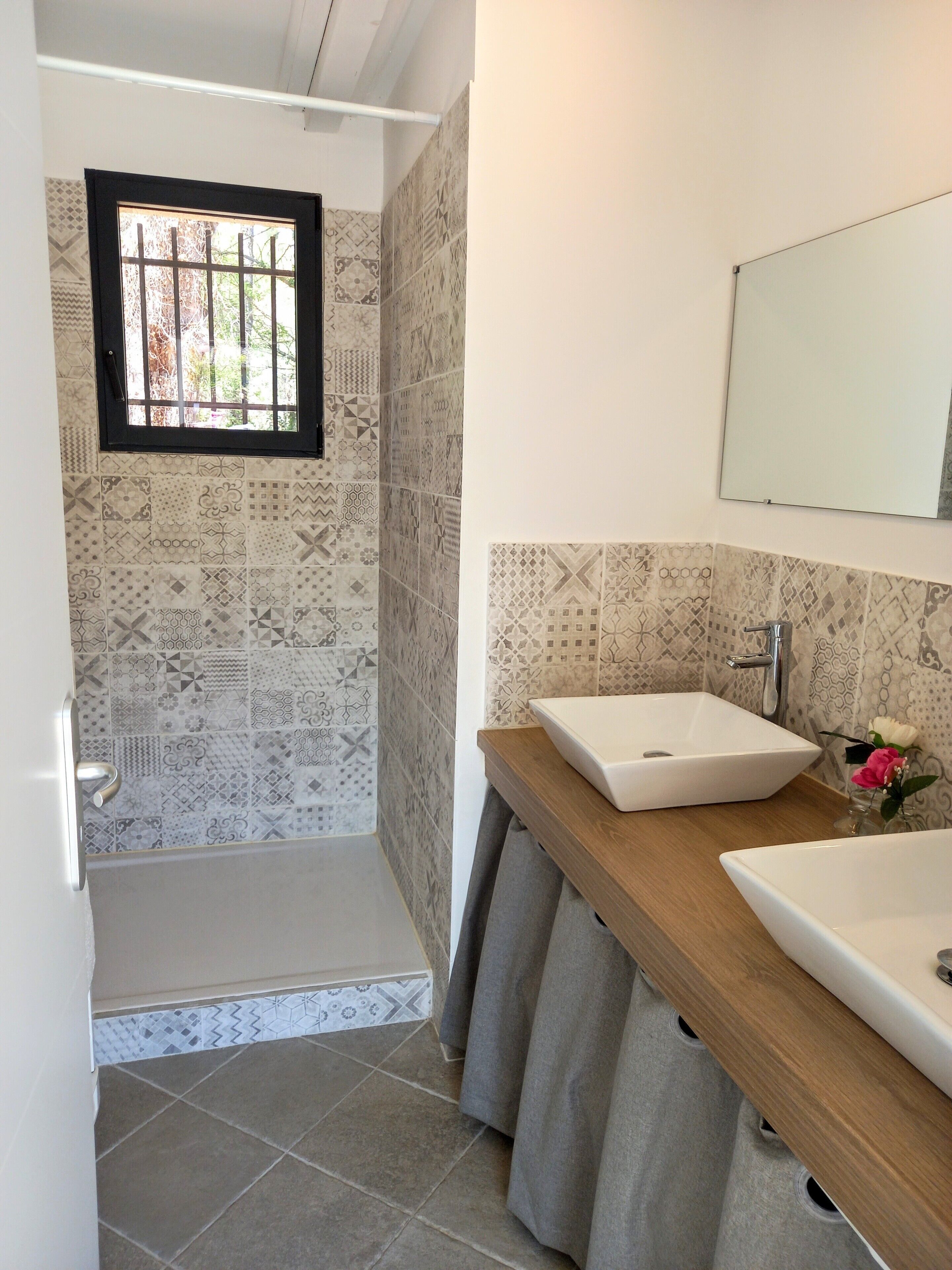 Une salle de bain avec des carreaux décorés, une fenêtre avec des barreaux, une douche, un lavabo et un miroir.