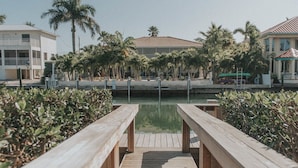 A wooden boardwalk leading to a calm water body, surrounded by palm trees and buildings with a Mediterranean architectural style. - Modern Island Luxury | 4 En-Suite Masters, 6 BA, Pool, Dock & Outdoor Oasis (Key Colony Beach)