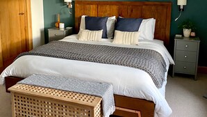 Une chambre à coucher avec un grand lit, une tête de lit en bois, un tableau représentant un paysage, une table de chevet, une lampe et une petite commode.