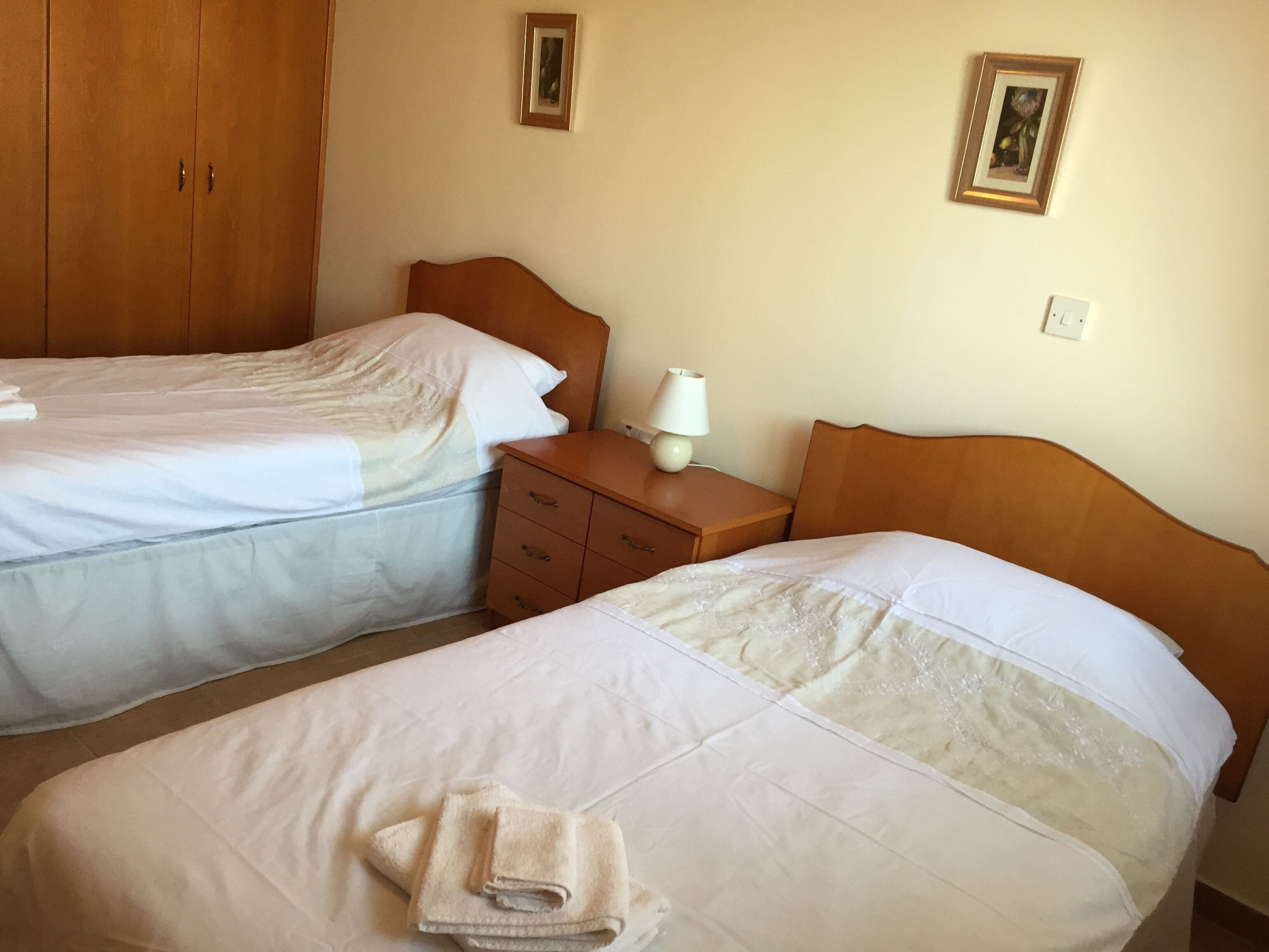 A hotel room with two beds, a wooden wardrobe, a nightstand with a lamp, and two framed pictures on the wall.