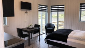 A modern room with a bed, a desk, and two chairs. There is a TV mounted on the wall and large windows with blinds. - Afternoon Cottages near Hella, Hekla & Landmannalaugar (Hella)