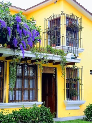 A two-story building with yellow walls, a balcony with purple flowers, and multiple windows with metal bars. - Villas Santa Ana-Ricardo (Antigua Guatemala)