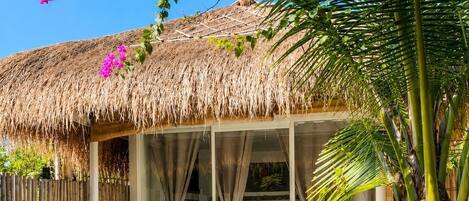 A thatched-roof outdoor area with a glass enclosure, a dining table, and a bed visible through the glass.