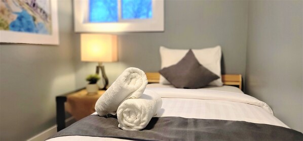 Una cama bien tendida con una almohada y una toalla enrollada en el borde. Una lámpara y una pequeña planta están sobre una mesita de noche.