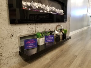 Un interior moderno con una televisión de pantalla plana montada en una pared con textura, una mesa consola negra con un cartel de prohibido fumar y una estantería decorativa con plantas y una escultura de metal.