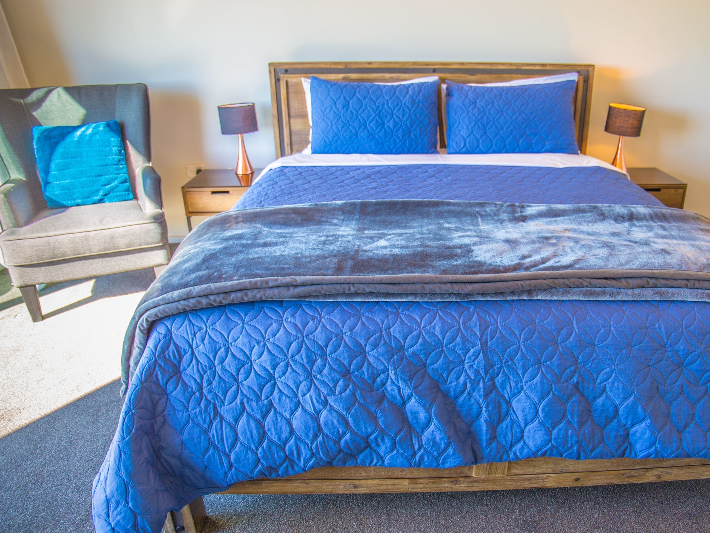 A bed with a blue quilted bedspread and pillows, flanked by two bedside tables with lamps, and a chair with a blue cushion.