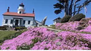 Un bâtiment en pierre blanche avec un toit rouge, un phare et des fleurs roses sur une colline.