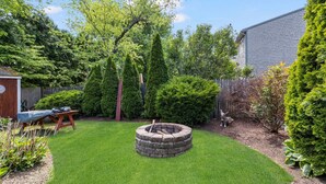 Ein Garten mit einem runden Feuerkorb aus Stein, einer Holzbank und verschiedenen Sträuchern und Bäumen.