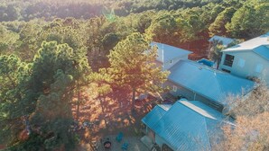 An aerial view of a house surrounded by dense forest, with a clear sky and sunlight filtering through the trees.