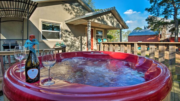 A hot tub with a bottle of wine and glasses, a wooden deck, and a house in the background.