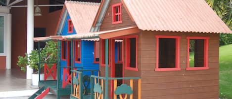 A children's playhouse with a red roof and blue walls, featuring a slide and a small porch.