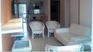 Een moderne woonkamer met een witte bank, twee witte fauteuils en een kleine tafel. Achtergrond toont een keuken met een magnetron en een eettafel.