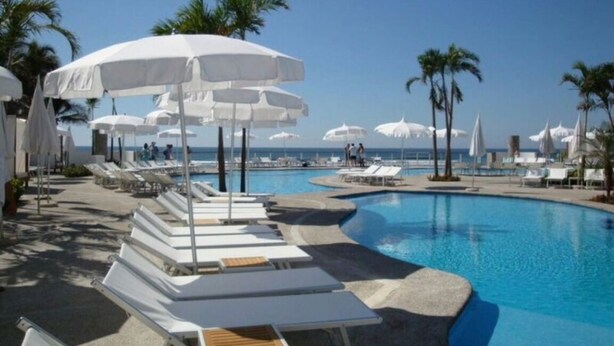 Un área junto a la piscina con sillones de descanso y sombrillas, palmeras y una vista despejada del océano.