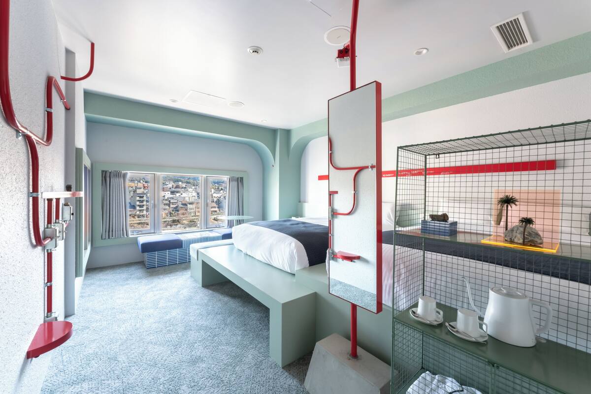 A modern bedroom with a bed, a bench, a large window, a mirror, and a small shelf with items.