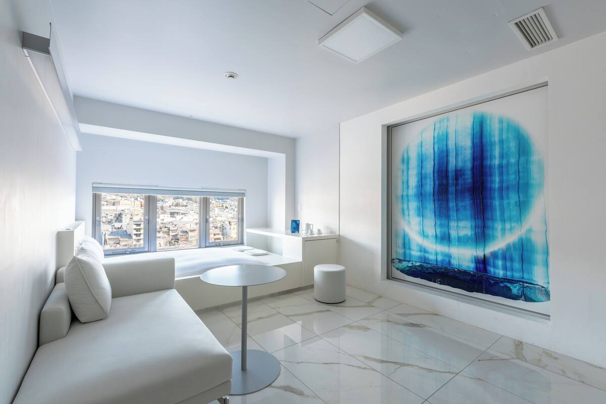 A modern hotel room with a large window, a white sofa, a round table, and a large abstract painting.
