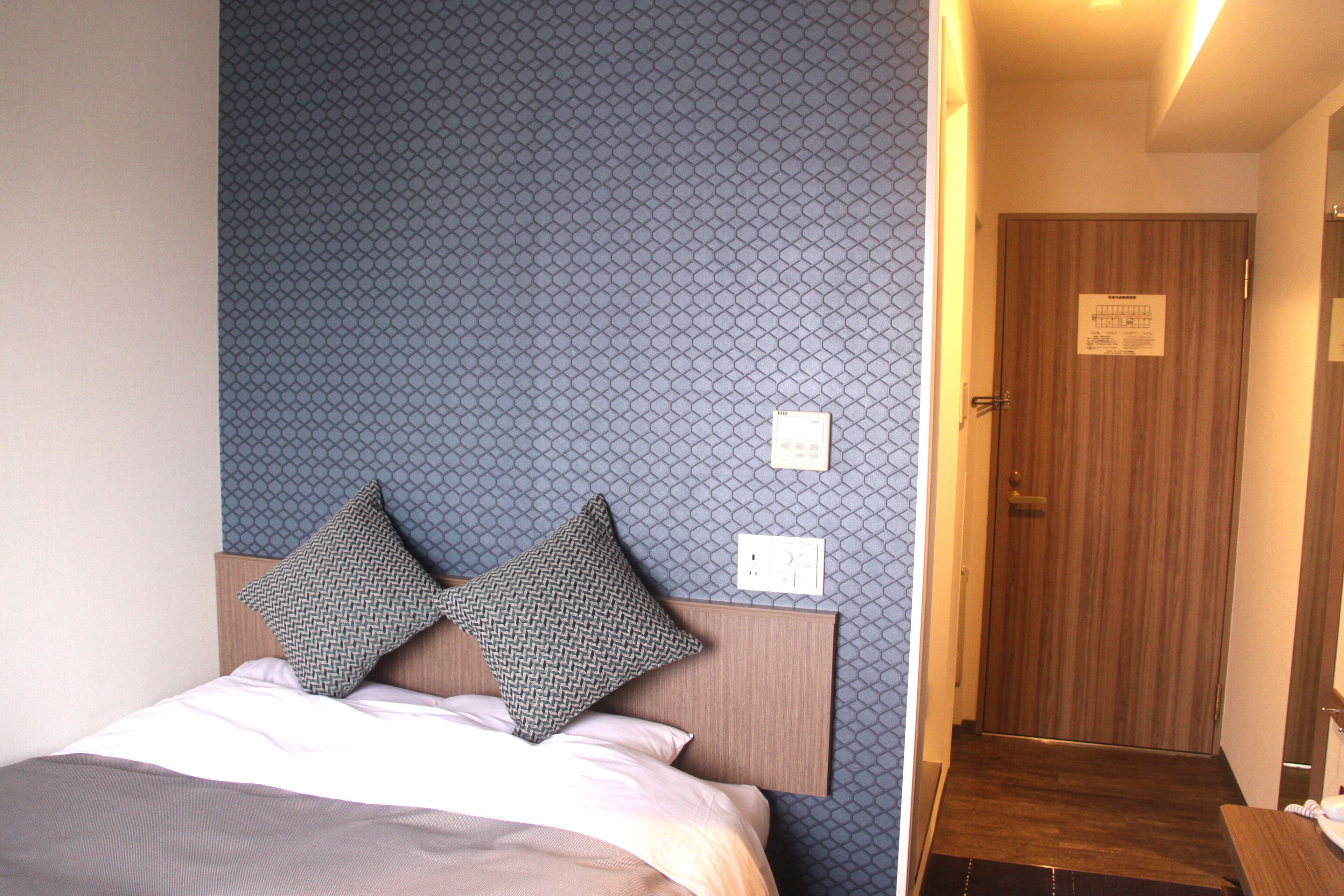 Habitación de hotel con cama, dos almohadas estampadas, cabecero, pared con diseño geométrico, puerta y un interruptor de luz.