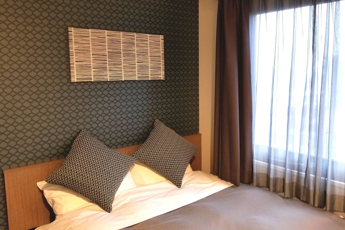 A hotel room with a bed, two patterned pillows, a headboard, a window with curtains, and a wall with a geometric pattern.