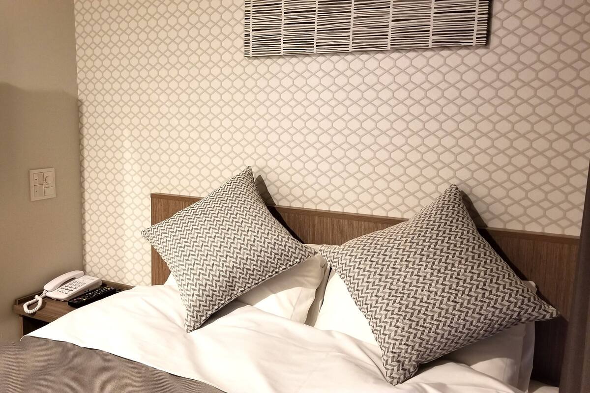 A hotel room with a bed, two patterned pillows, a bedside telephone, and a wall-mounted air conditioning unit.