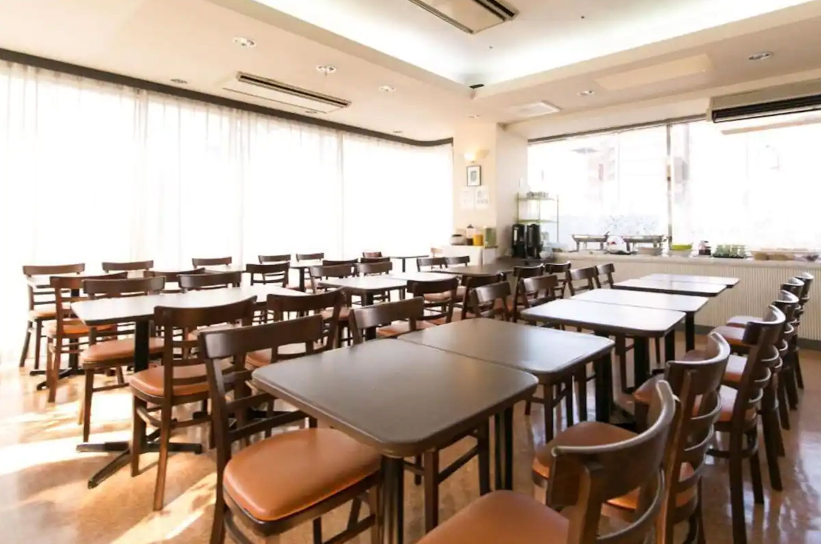 A spacious dining area with multiple tables and chairs, large windows with curtains, and a kitchen area in the background.