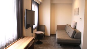 Habitación de hotel con sofá, una mesita redonda, televisor montado en la pared y ventana con cortinas.