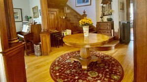 A spacious living room with a high ceiling, wooden beams, a round coffee table with a floral rug, a chandelier, and a staircase leading to an upper level.