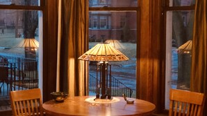 A corner dining area with a round wooden table, two wooden chairs, a table lamp, and a view of a house through large windows with curtains.