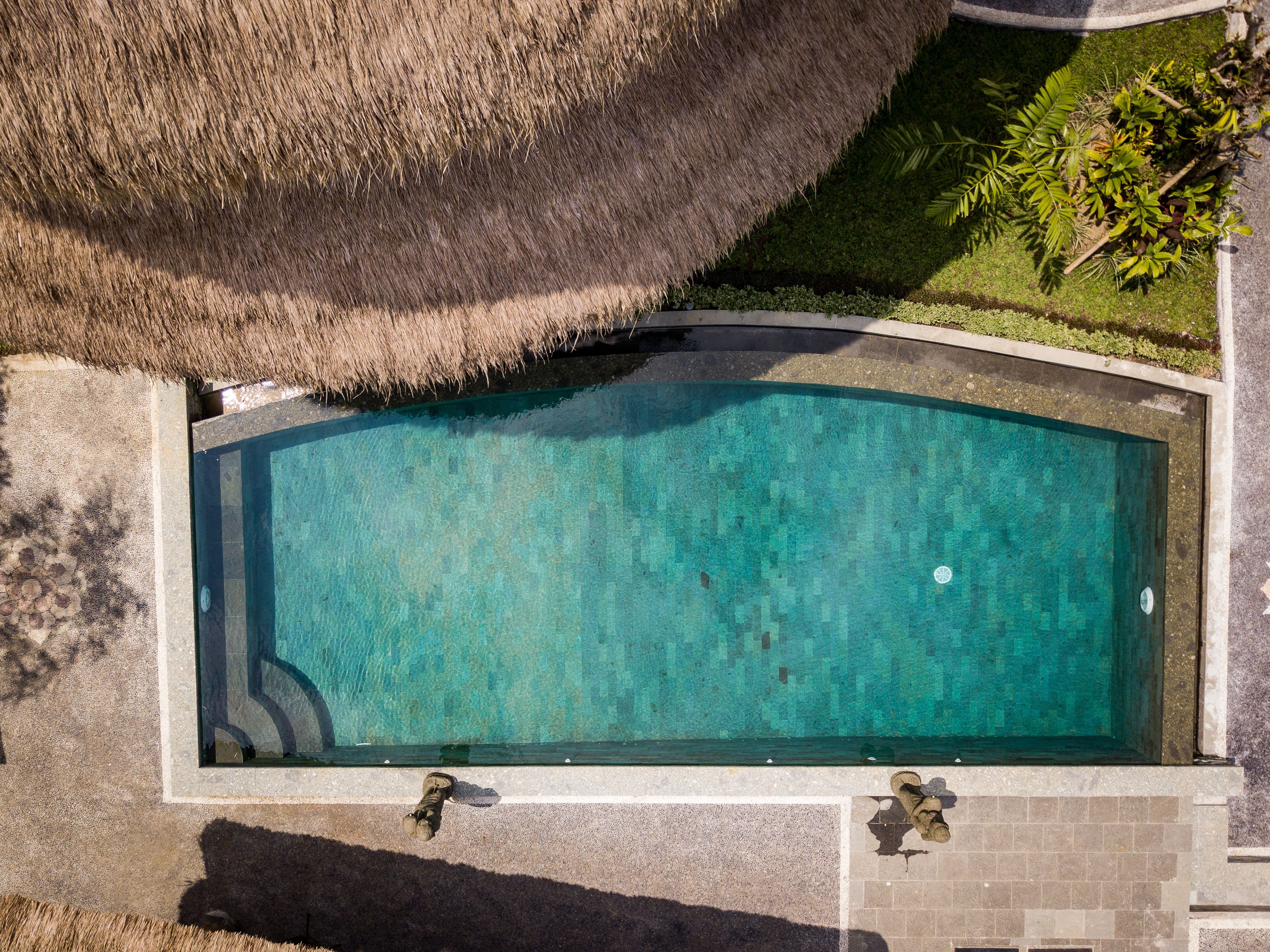 Vista aerea di una piscina rettangolare con una struttura a tetto di paglia su un lato, circondata da pavimentazione e vegetazione.
