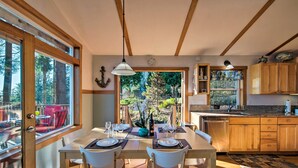 Un coin repas avec une table en bois dressée pour deux personnes, une vue sur l’extérieur grâce à de grandes fenêtres, et une cuisine dotée de meubles en bois.