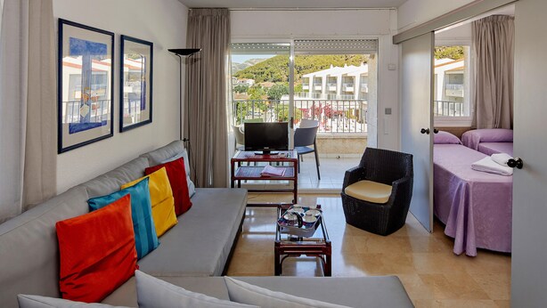 Una sala de estar moderna con un sofá, una televisión y un balcón con vistas a edificios y colinas.