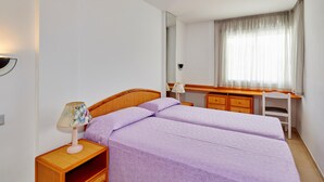 A bedroom with two single beds, a wooden nightstand with a lamp, a desk, and a chair.
