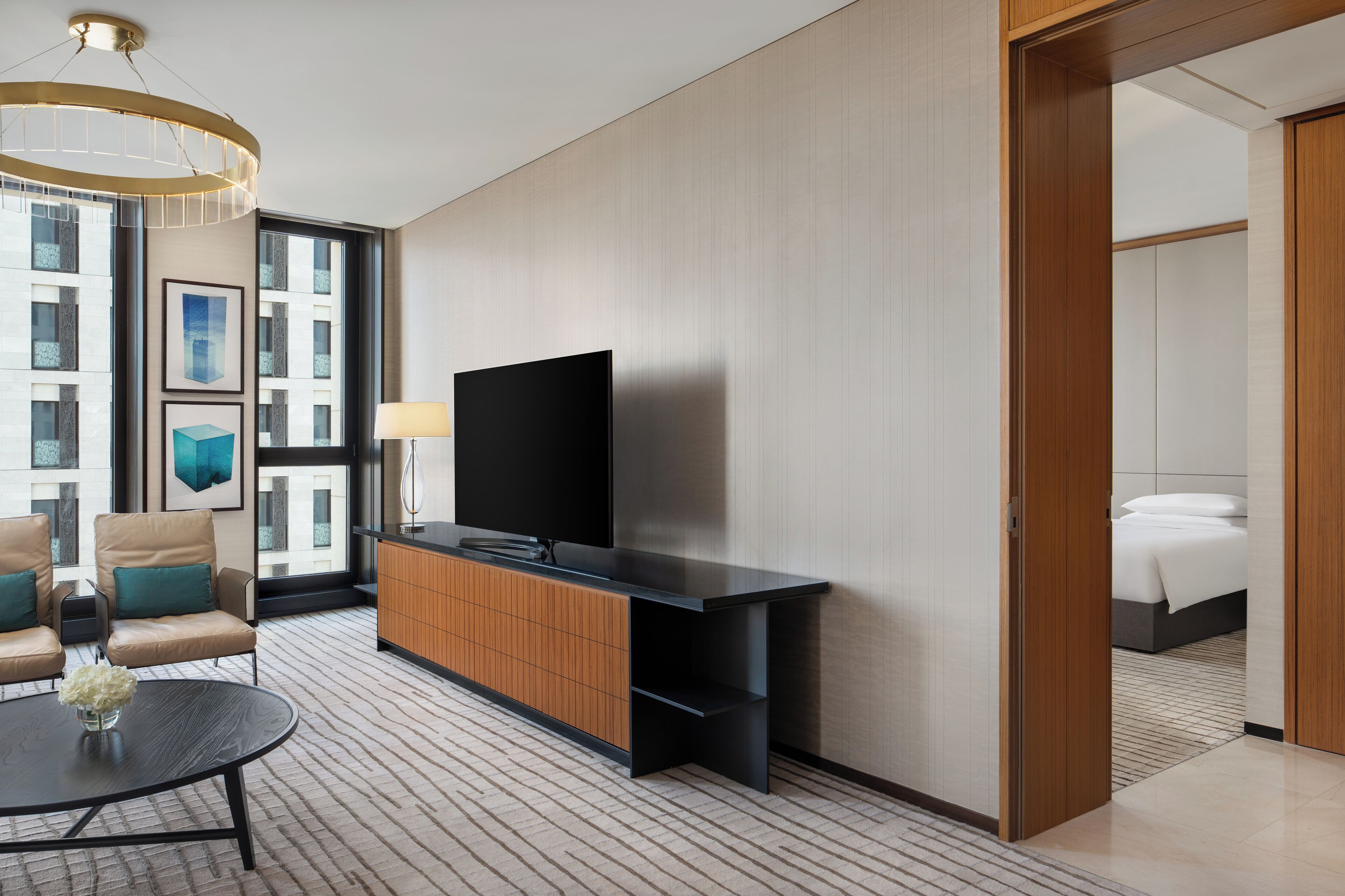 Habitación de hotel moderna con un televisor de pantalla plana grande, una mesa consola de madera, una mesa redonda de centro con un florero y un área de descanso con sillones de cuero.