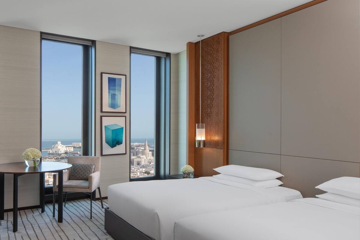 Ein Hotelzimmer mit zwei Betten, einem Schreibtisch, einem Stuhl und einem großen Fenster mit Blick auf die Stadt.