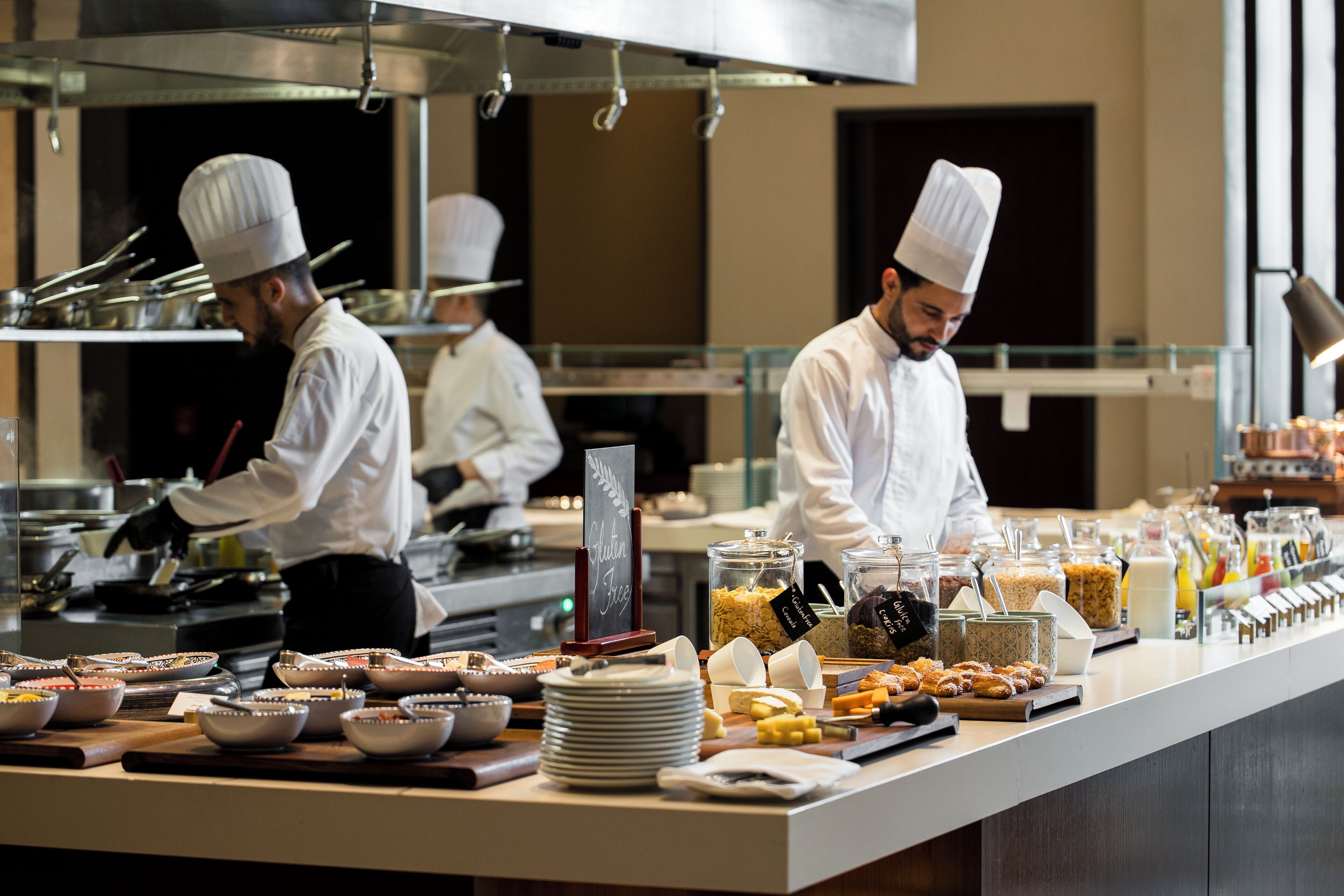 Montaje de bufé con diversos alimentos y chefs preparando comida en una cocina profesional.