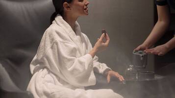 Una persona con bata blanca sentada en una silla negra en una habitación con poca luz y una ducha de vapor.