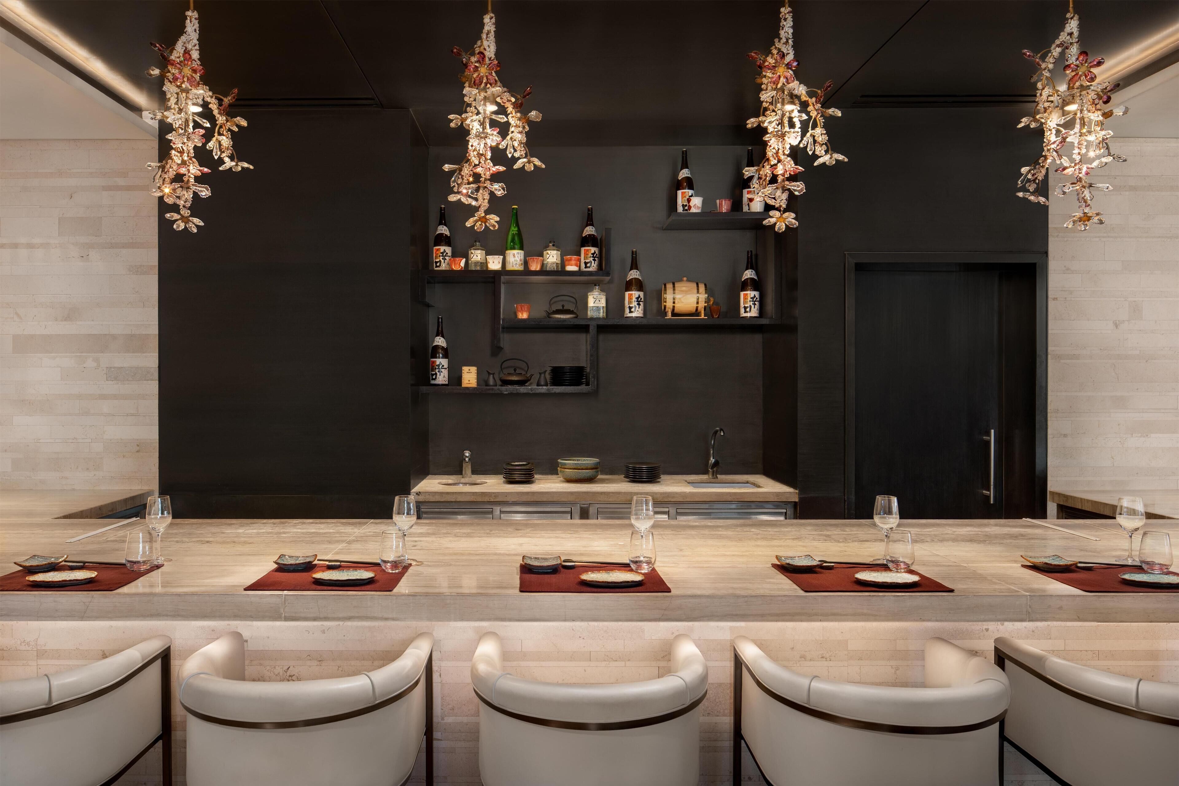 Un comedor moderno con una larga mesa dispuesta para cuatro personas, rodeada de sillas blancas. Sobre la mesa, hay lámparas decorativas colgantes. En el fondo, se observan estanterías con botellas y objetos decorativos.