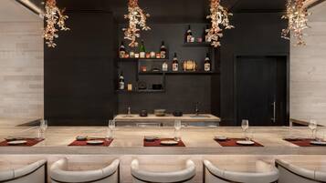 Un comedor moderno con una larga mesa dispuesta para cuatro personas, rodeada de sillas blancas. Sobre la mesa, hay lámparas decorativas colgantes. En el fondo, se observan estanterías con botellas y objetos decorativos.