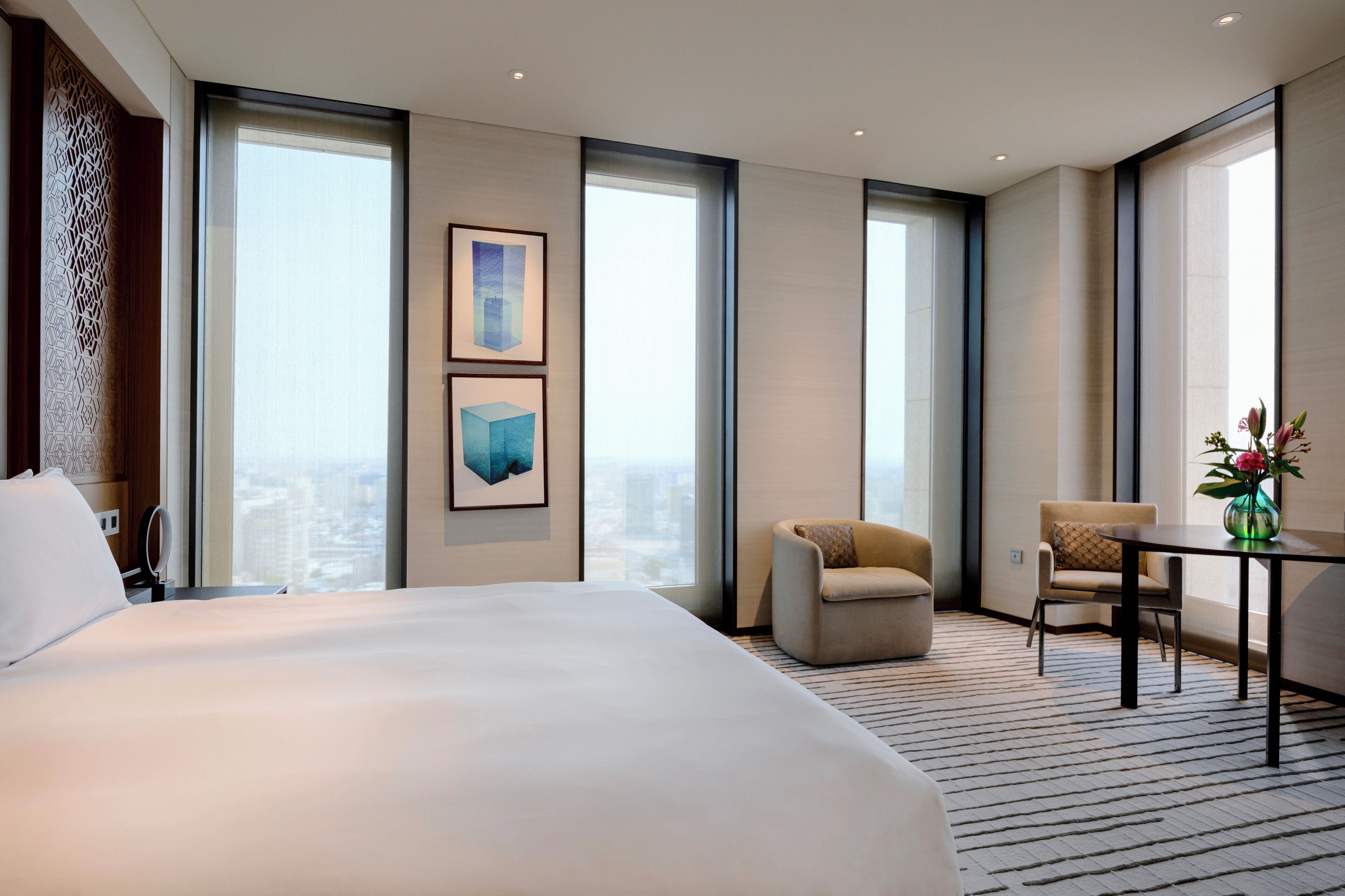 A hotel room with a large bed, a sofa, a small table with a vase, and large windows offering a city view.