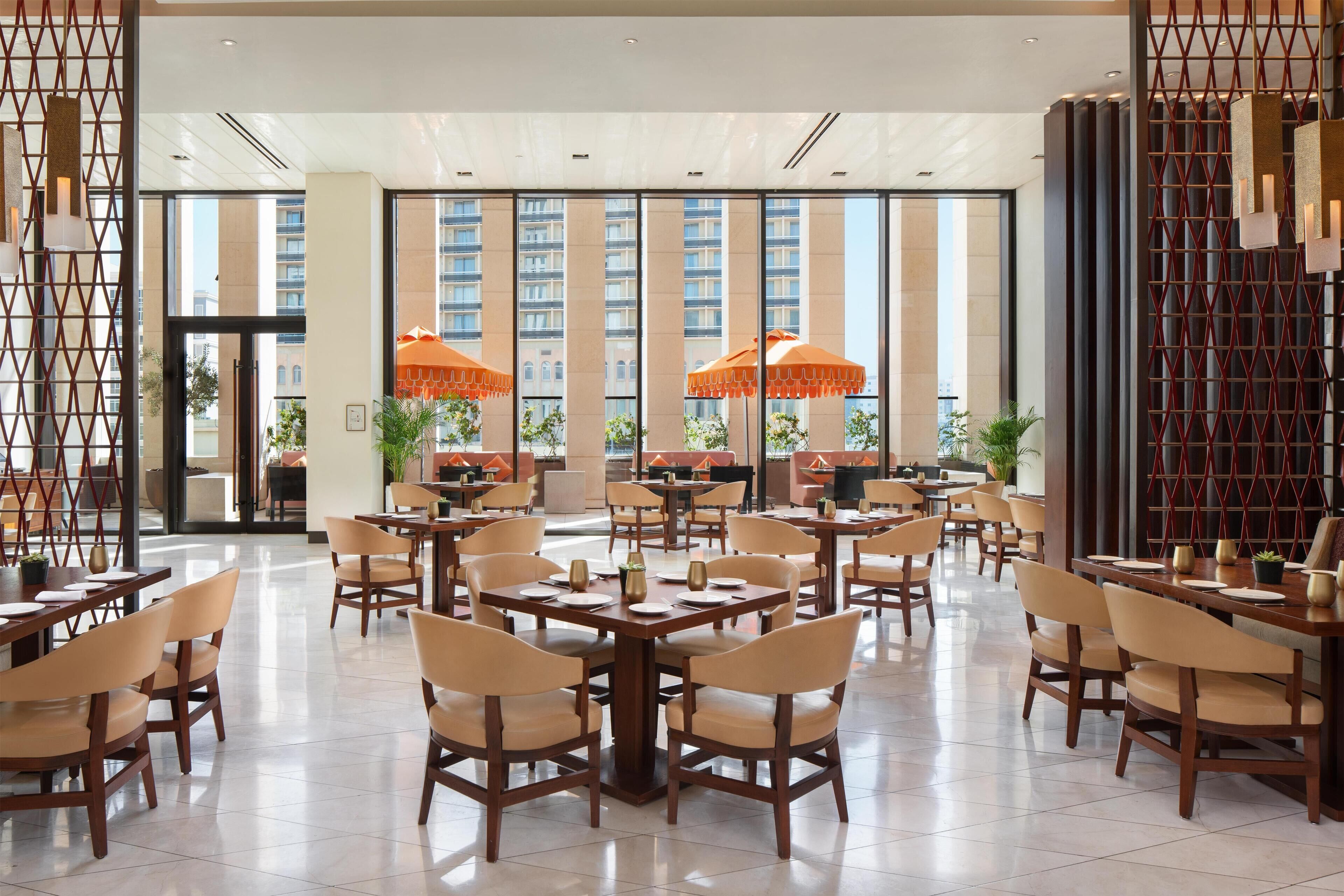 A spacious indoor dining area with multiple tables and chairs, large windows, and orange umbrellas.