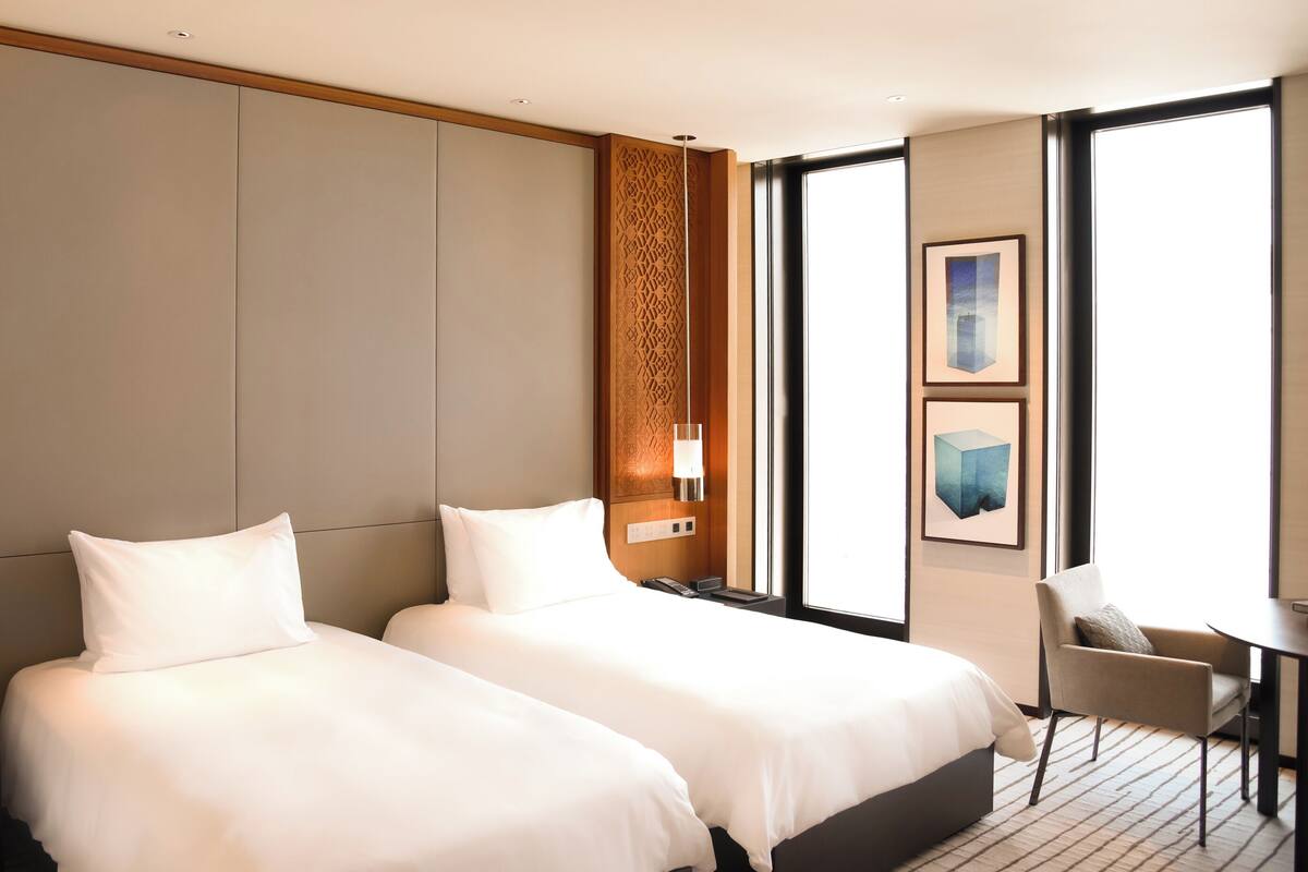 Ein Hotelzimmer mit zwei Betten, einem Schreibtisch, einem Stuhl und großen Fenstern.