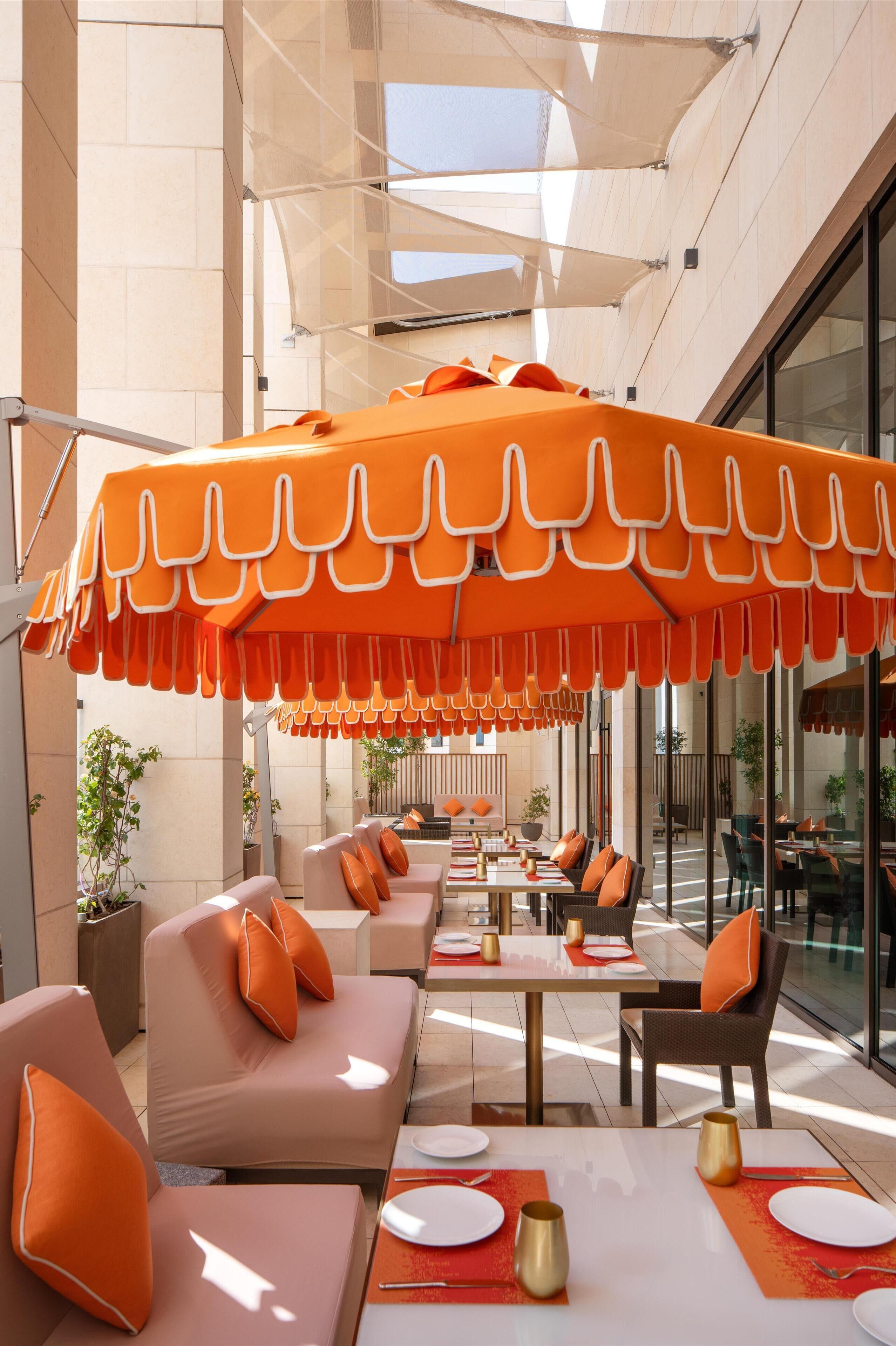 Un área de comedor exterior moderna con sombrillas naranjas, asientos acolchados y una pared de vidrio que ofrece vistas a la calle.