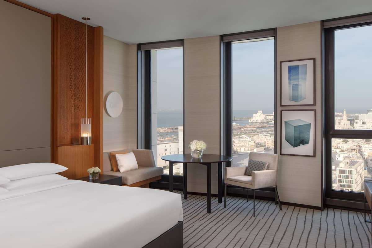 Ein Hotelzimmer mit einem großen Bett, einer Couch, einem kleinen Tisch und Blick auf die Stadt durch große Fenster.