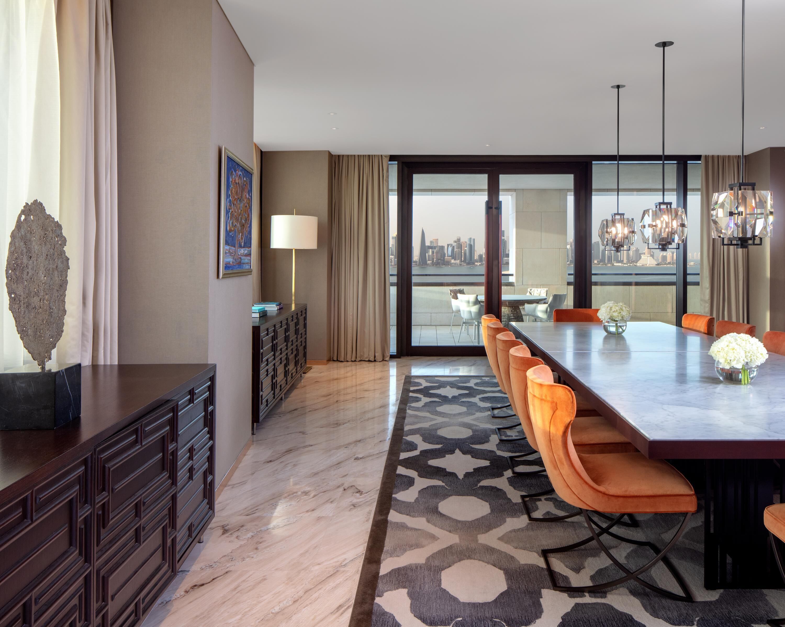 A modern hotel dining area with a long table, orange chairs, a patterned rug, and a city view through large windows.