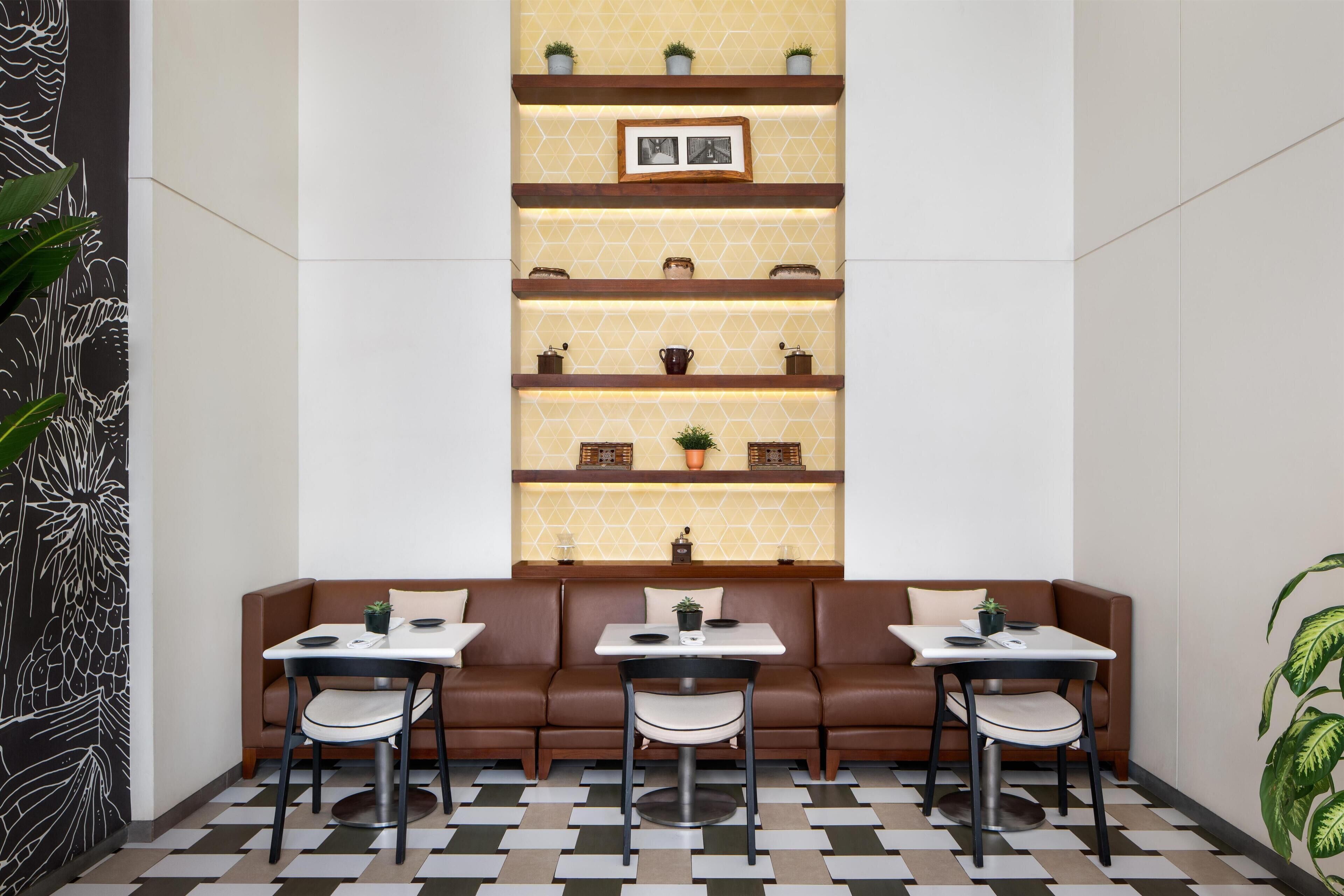 Un intérieur moderne avec un ensemble de canapés marron, deux tables et des chaises, un sol à motifs géométriques et des étagères ornées d’objets décoratifs.