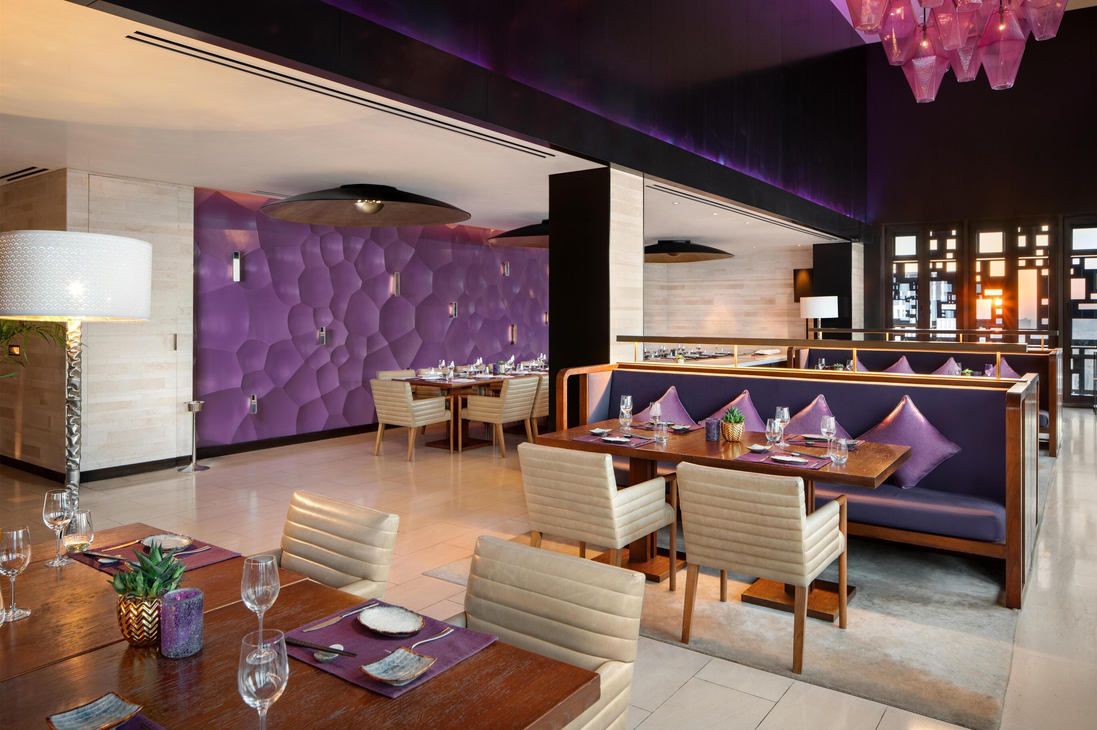 A modern dining area with a purple textured wall, wooden tables, and cushioned seating.